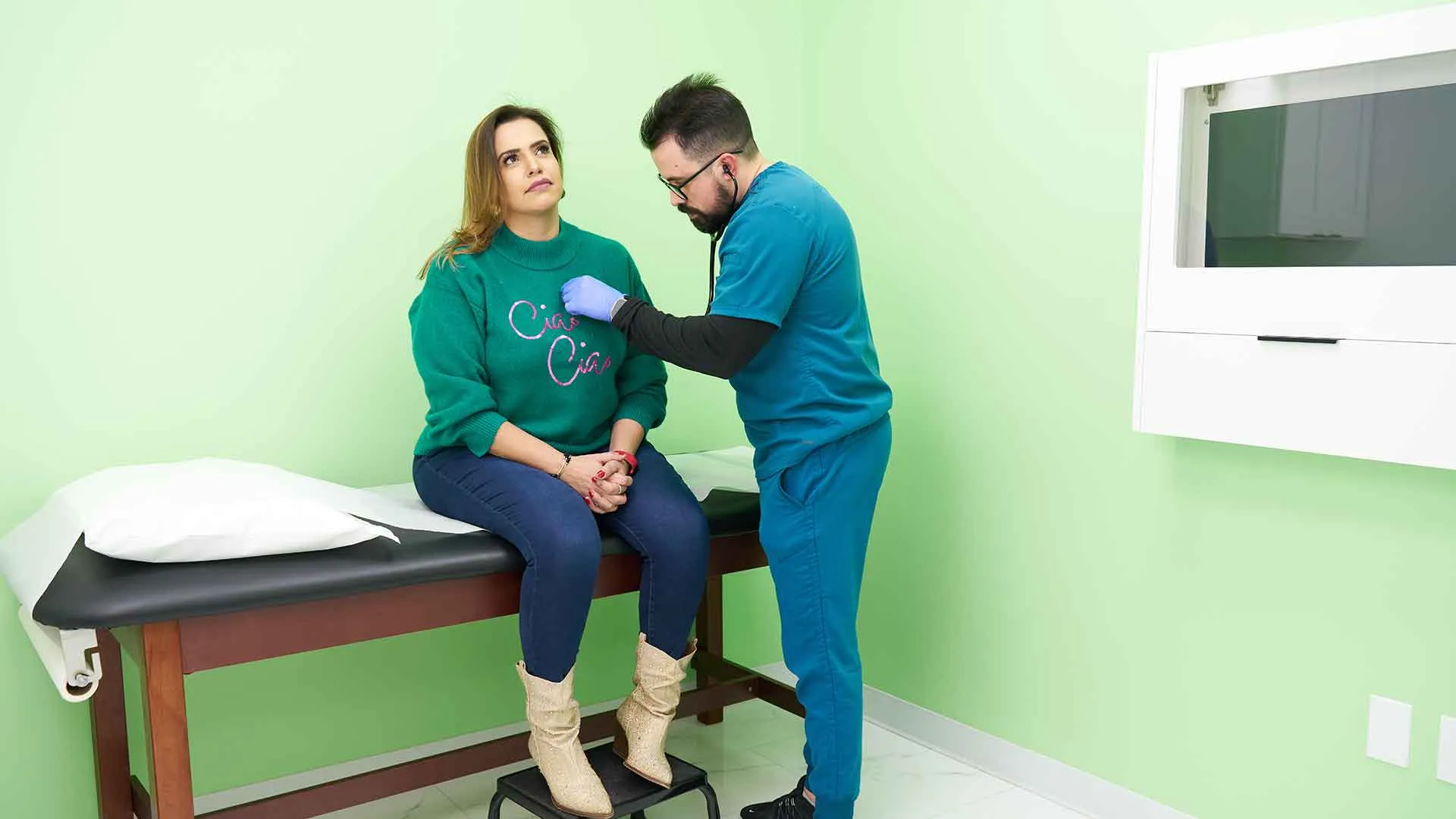 Médico con uniforme azul y guantes examina a la paciente sentada en la camilla dentro del consultorio de la Clínica la Virgen de Guadalupe