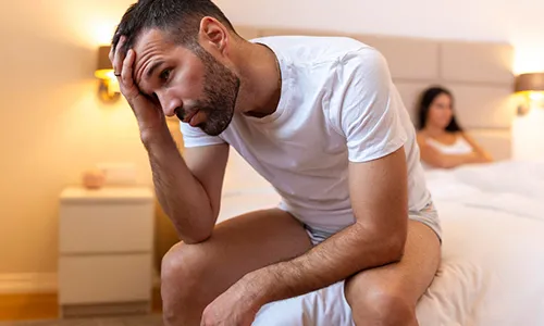 Hombre joven sentado al borde de la cama con expresión de cansancio y dolor de cabeza, sosteniendo su frente.